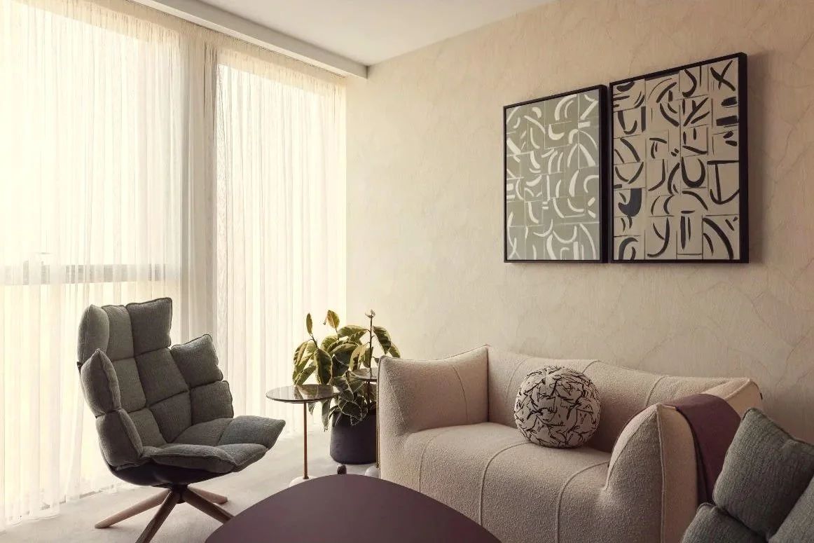 Living room with a gray armchair, a plant, a cream-colored sofa with a patterned round pillow, and abstract artwork on the wall, illuminated by natural light from large curtains.