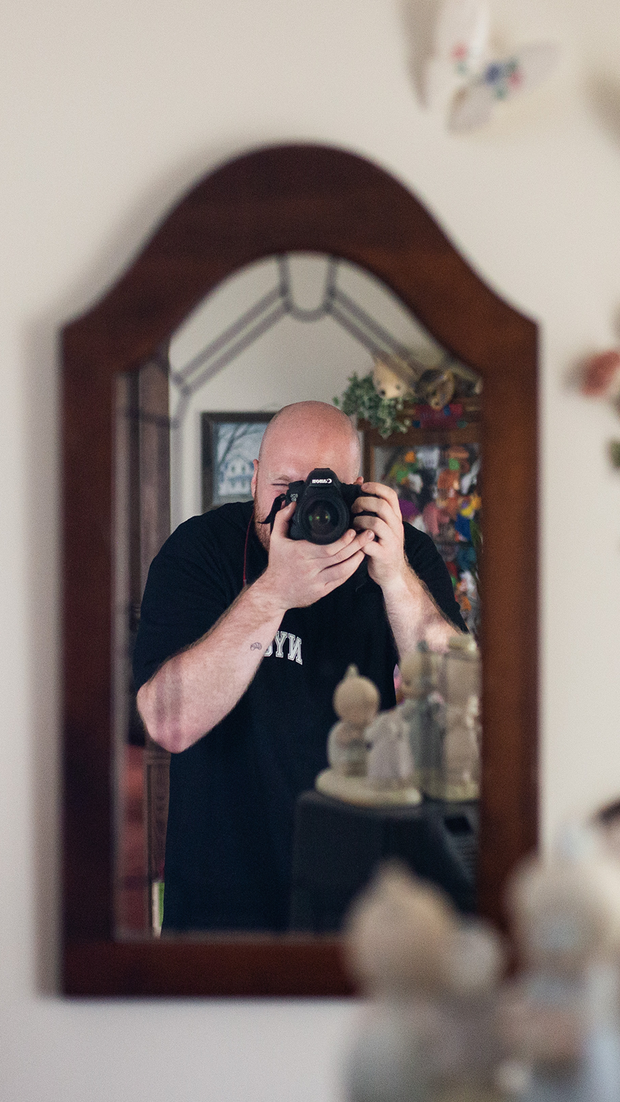 A person taking a photo of their reflection in a mirror with a DSLR camera, showing a room with some decor and framed pictures in the background.