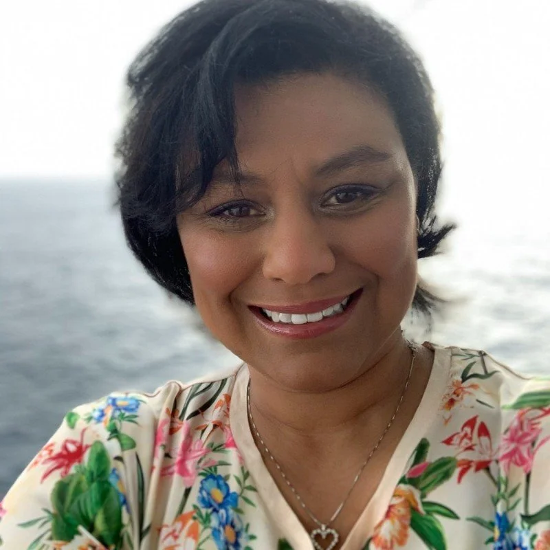 A woman smiling outdoors with water in the background, wearing a floral top and a heart-shaped necklace.