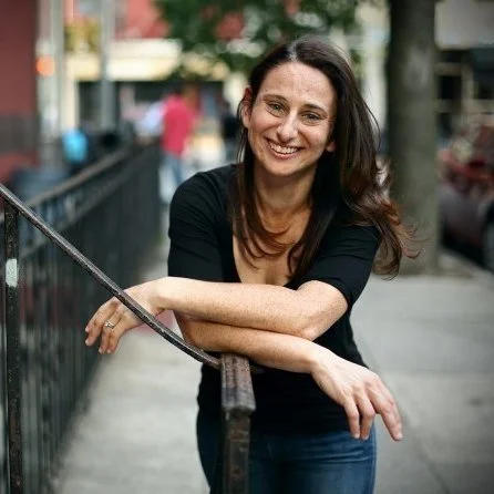 Smiling woman with long dark hair in a black shirt leaning on railing on a sidewalk in an urban area.
