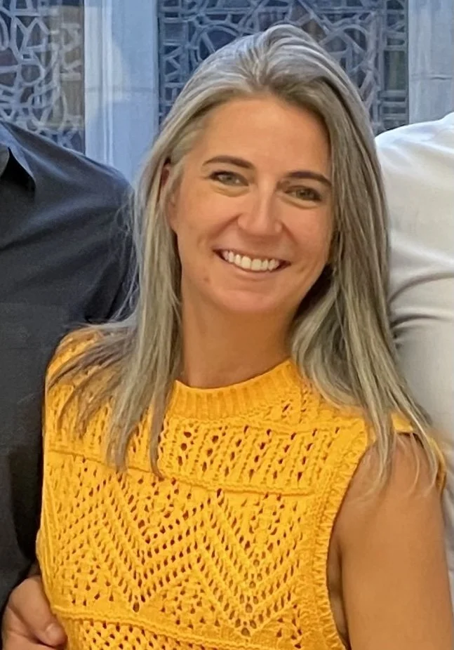 Smiling woman with long gray hair wearing a yellow crochet top, standing indoors with windows in the background.