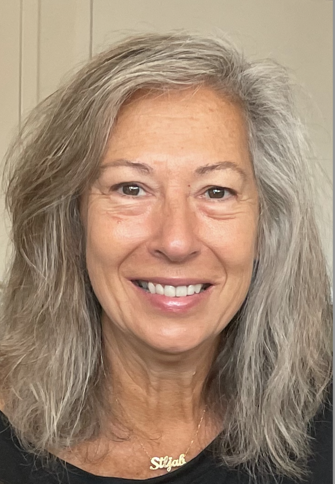 A woman with grey, shoulder-length hair, smiling, wearing a black top and gold necklace with the name "Siljak" in cursive.