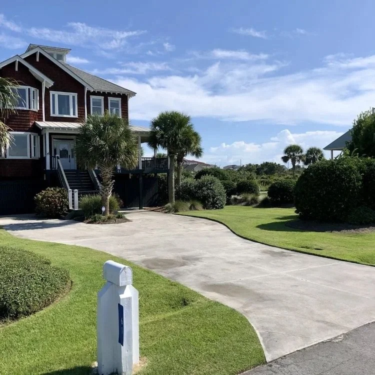A large house with a wooden exterior, multiple windows, and stairs leading to the entrance, surrounded by palm trees, bushes, and a well-maintained lawn, with a driveway and a mailbox in the foreground.