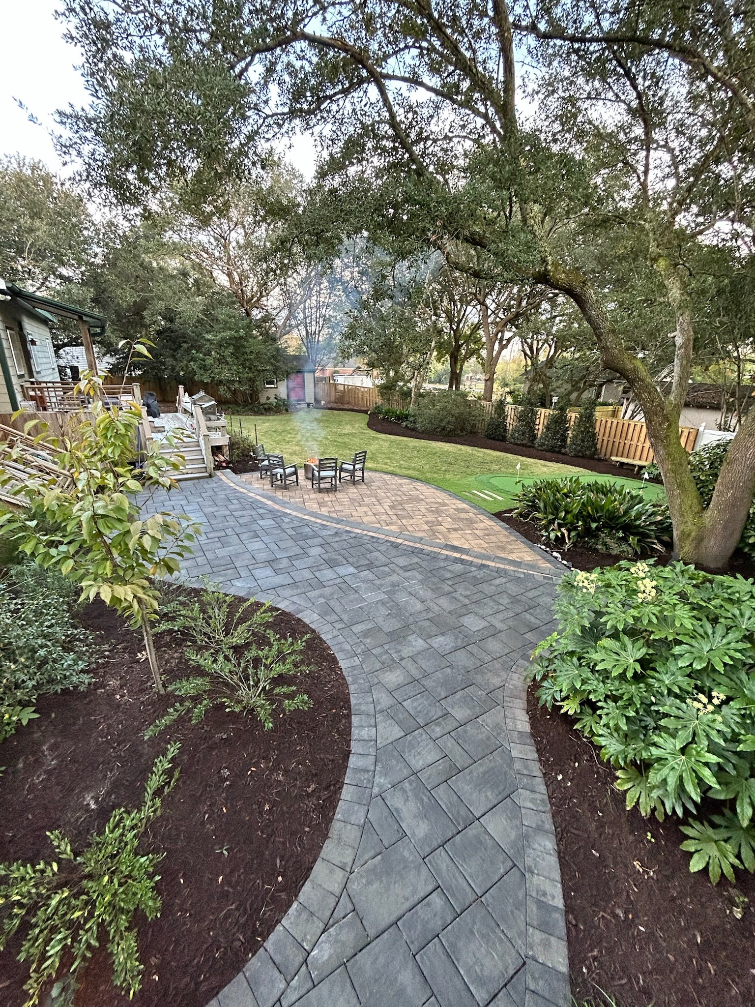 A backyard patio with a curved paving stone pathway, outdoor seating around a fire pit, surrounded by plants, trees, and a wooden fence.