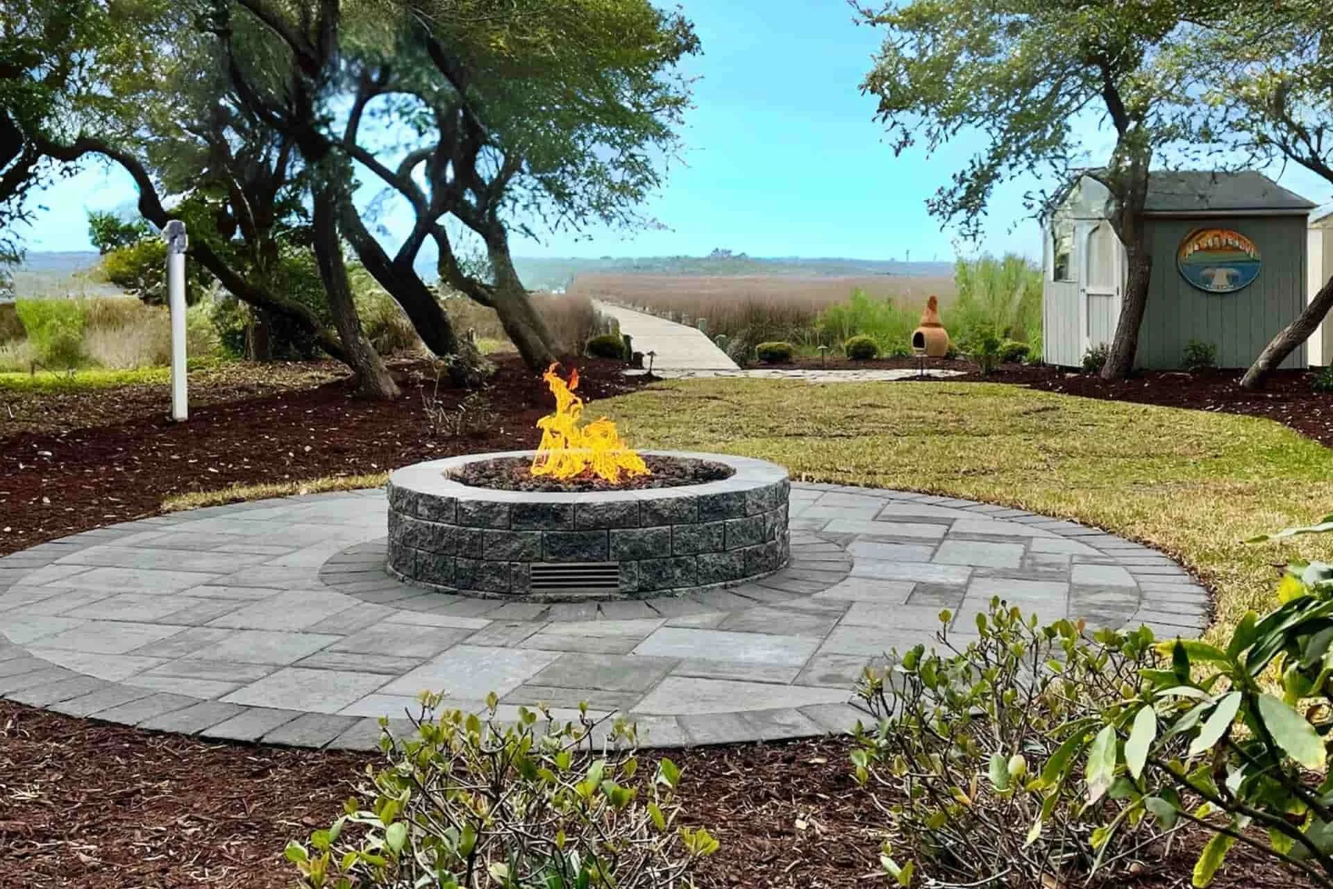 A backyard patio with a circular fire pit, surrounded by gray stone pavers, and trees providing shade. A pathway leads to a distant open field with a blue sky.