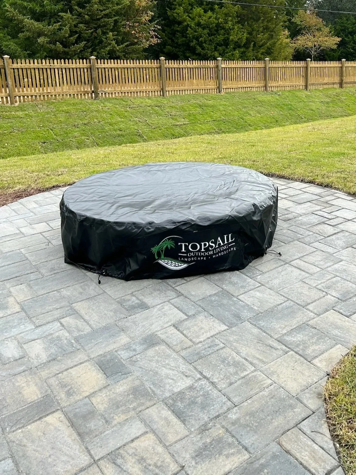 A covered round hot tub on a gray brick patio, with a wooden fence and green trees in the background.