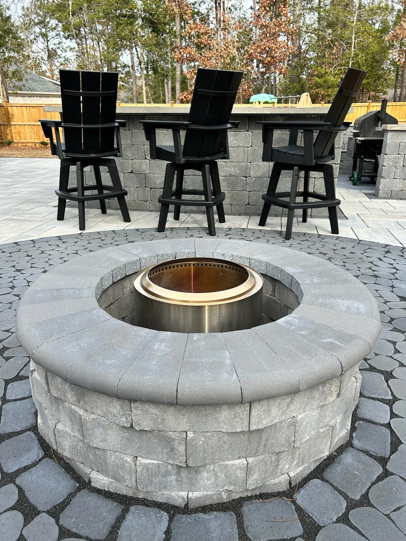 A circular stone fire pit with a metal fire insert in an outdoor patio, surrounded by gray paving stones and a wooden fence. Three black Adirondack chairs are positioned behind the fire pit.