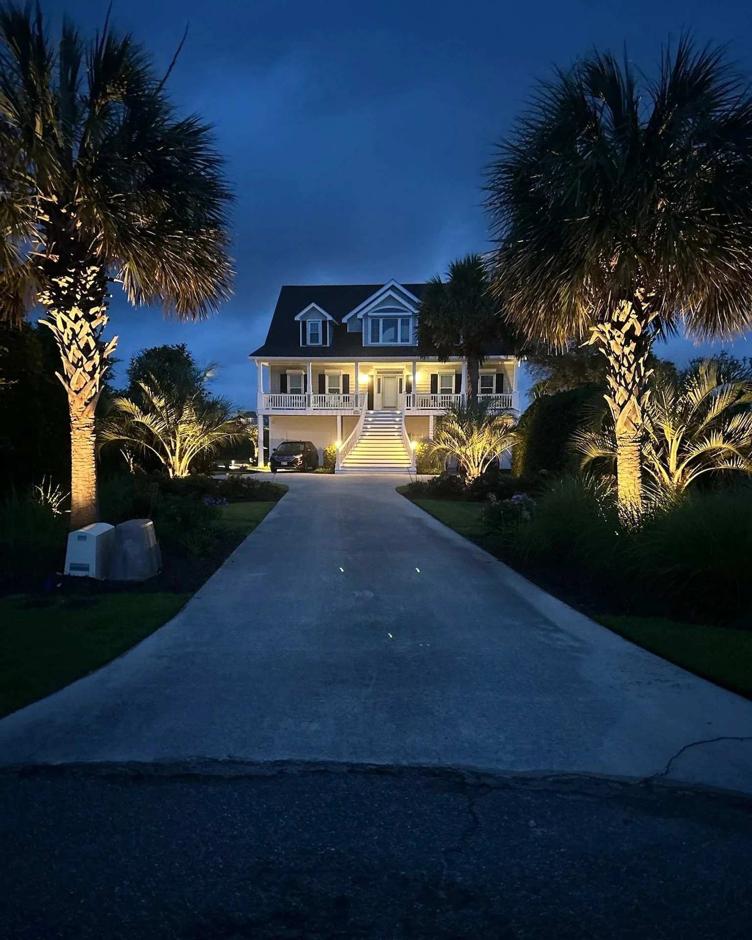 Nighttime view of a house with a porch and staircase, surrounded by palm trees with lights, parked cars, and a driveway, under a dark blue sky.