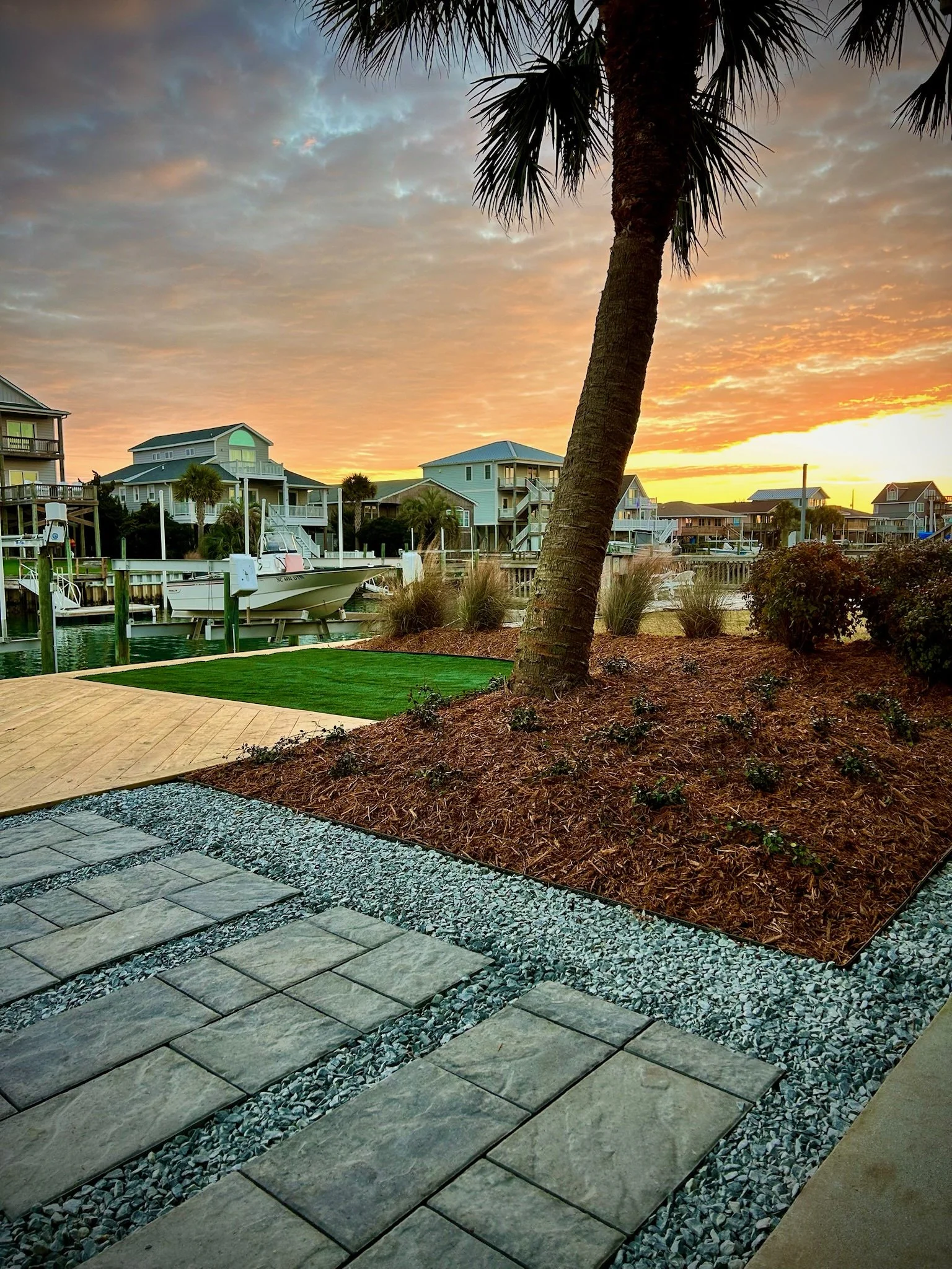 A waterfront residential area during sunset with houses on stilts, a boat docked in the water, a large palm tree, and a paved patio with a landscaped garden in the foreground.