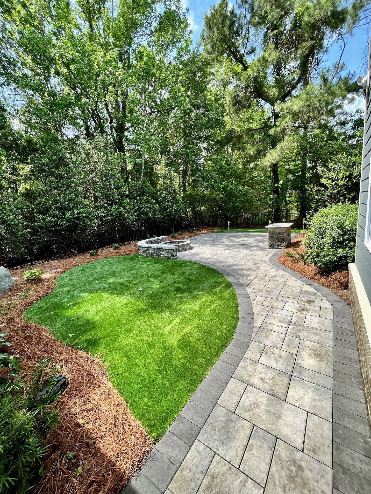 A landscaped backyard with a curved stone pathway, a small circular fire pit, a well-manicured green lawn, and trees and bushes in the background.