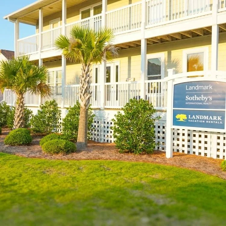 A two-story yellow vacation rental house with a white balcony railing, surrounded by palm trees and shrubs, with a turf lawn in the foreground and a sign for Landmark Sotheby's and Landmark Vacation Rentals.
