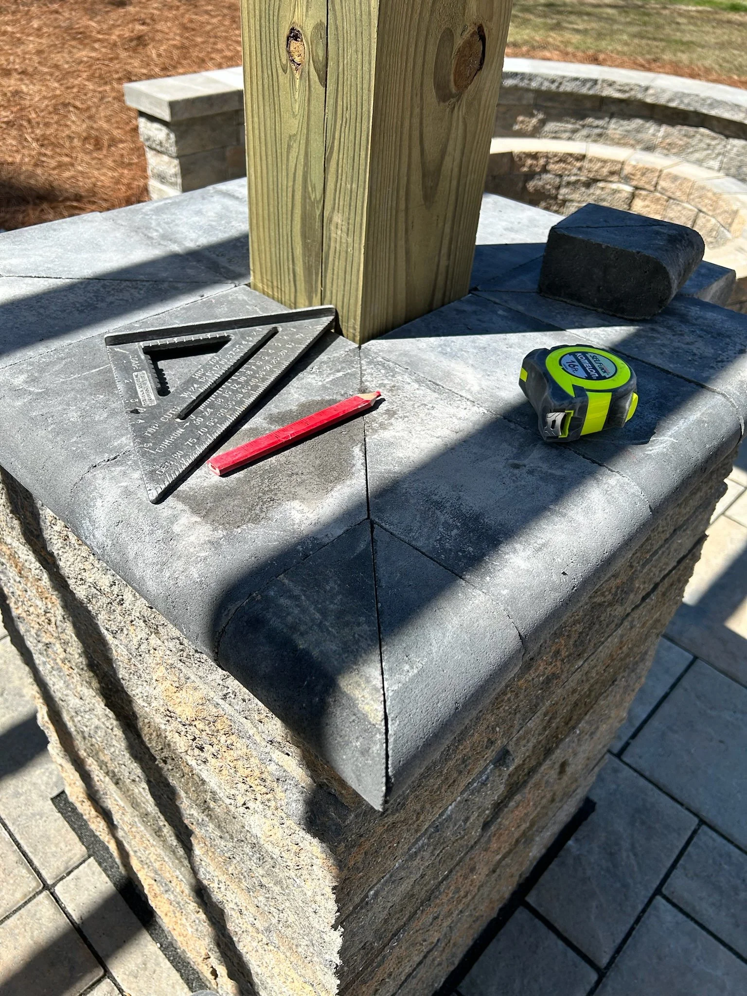 A close-up of a construction site showing a stone column with a wooden post, a level measuring tape, a red pencil, a carpenter's square, and a black stone on top of the pedestal.