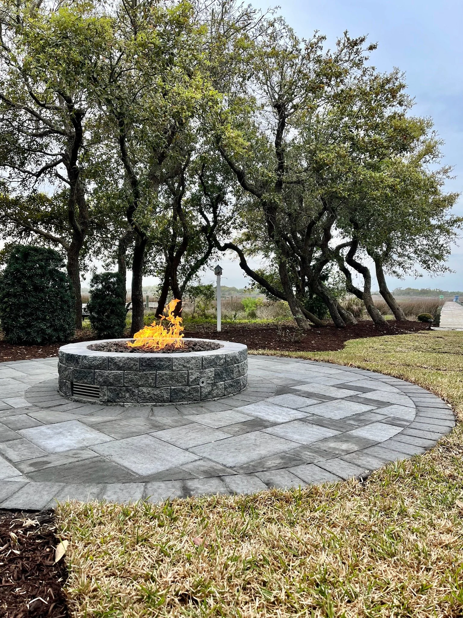 A stone fire pit with a small flame on a circular stone patio with grass and trees in the background.