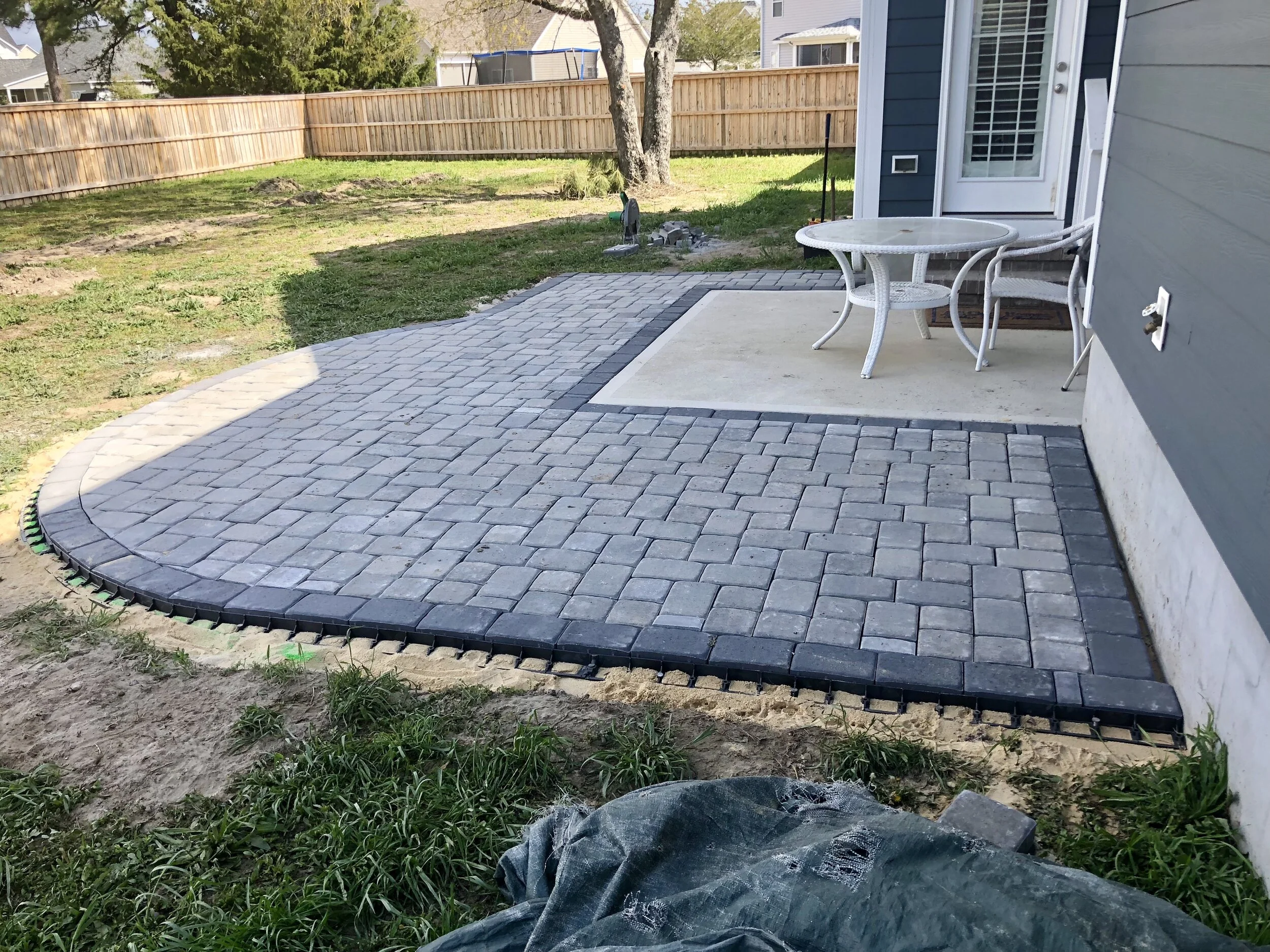 A backyard patio with fresh gray brick pavers and a small round gray table with white chairs, surrounded by a wooden fence and green grass, with a house wall on the right.