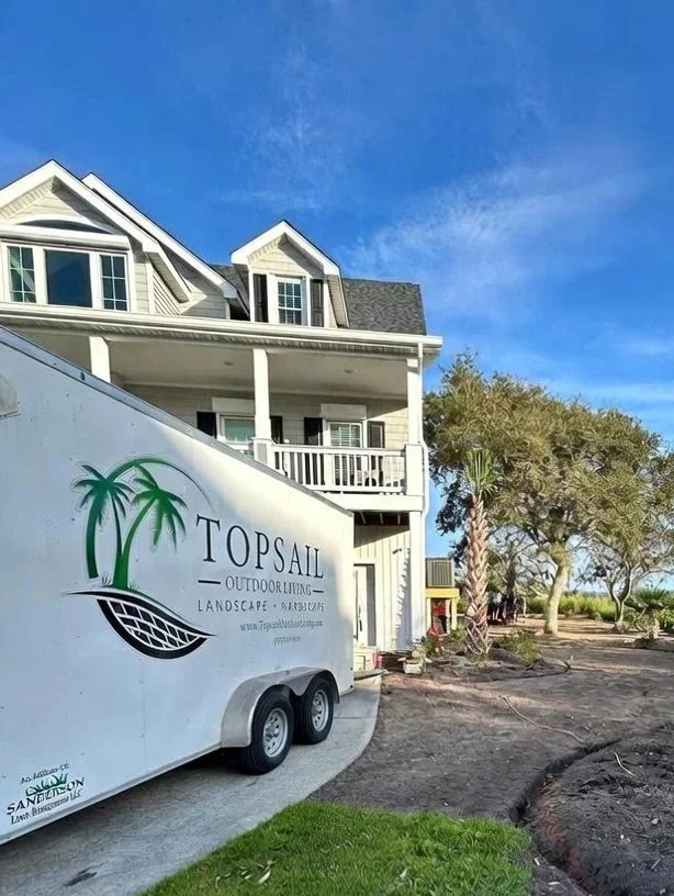 A white trailer labeled 'Topsail Outdoor Living Landscape & Arborist' parked on a curved driveway next to a large, multi-story house with a balcony, under a blue sky with some clouds, and surrounded by trees and landscaping work.