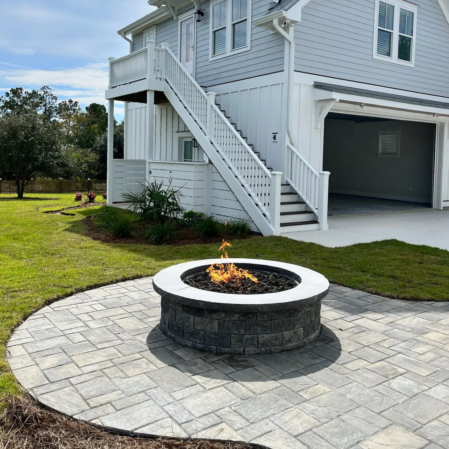 Backyard with a fire pit on a paved circular patio, a fenced lawn, and a white house with stairs leading to a second floor balcony.