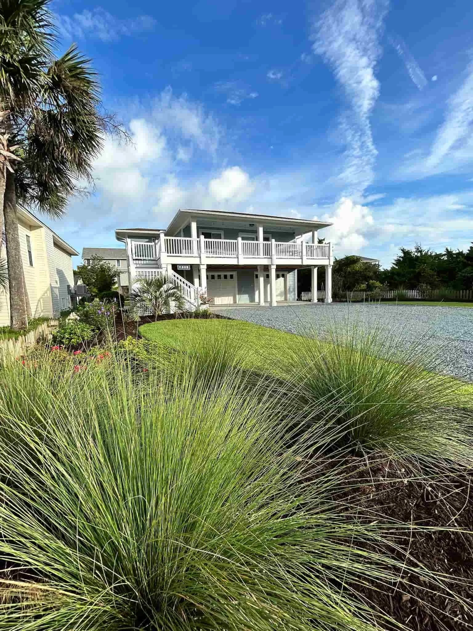 A house with a large front porch and white balcony, situated in a landscaped yard with lush green grass, spiky ornamental grass in the foreground, and a partly cloudy blue sky overhead.