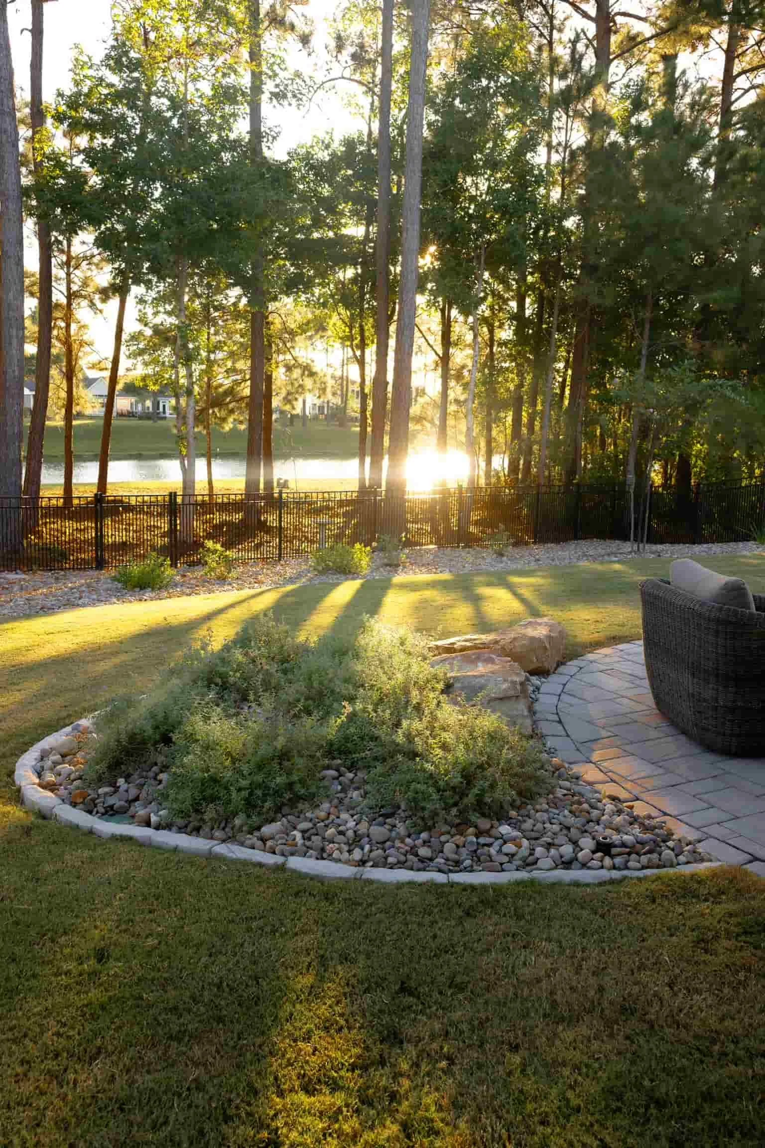 A backyard with a well-manicured lawn, a small pond, and a raised flower bed with rocks and greenery. In the background, tall trees are lit by the setting sun, creating a warm glow. There is a black metal fence separating the yard from the pond, and a patio area with a curved brick border and outdoor furniture.
