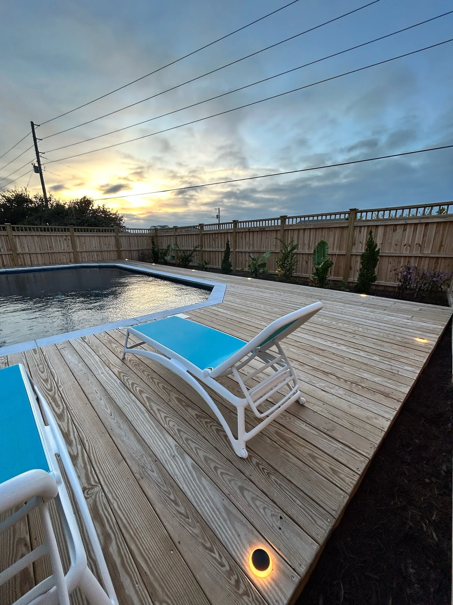 Wooden deck with a pool, blue lounge chairs, a wooden fence, small plants, and a sunset sky with clouds.