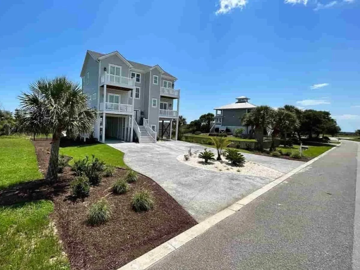 A coastal residential neighborhood with elevated beach houses, including a gray house with multiple balconies and stairs, surrounded by palm trees and landscaped yard, under a bright blue sky.