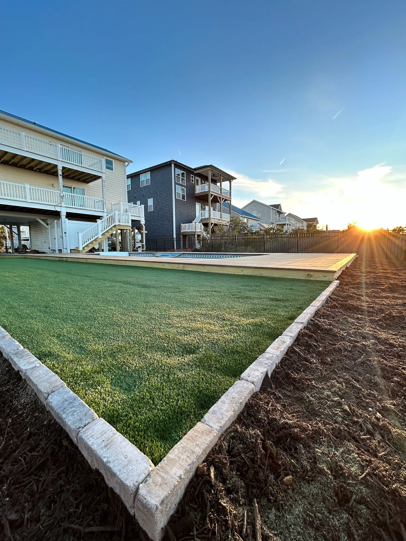 A row of newly laid green artificial grass lawns with white stone borders in a residential area, with houses and a sunset in the background.