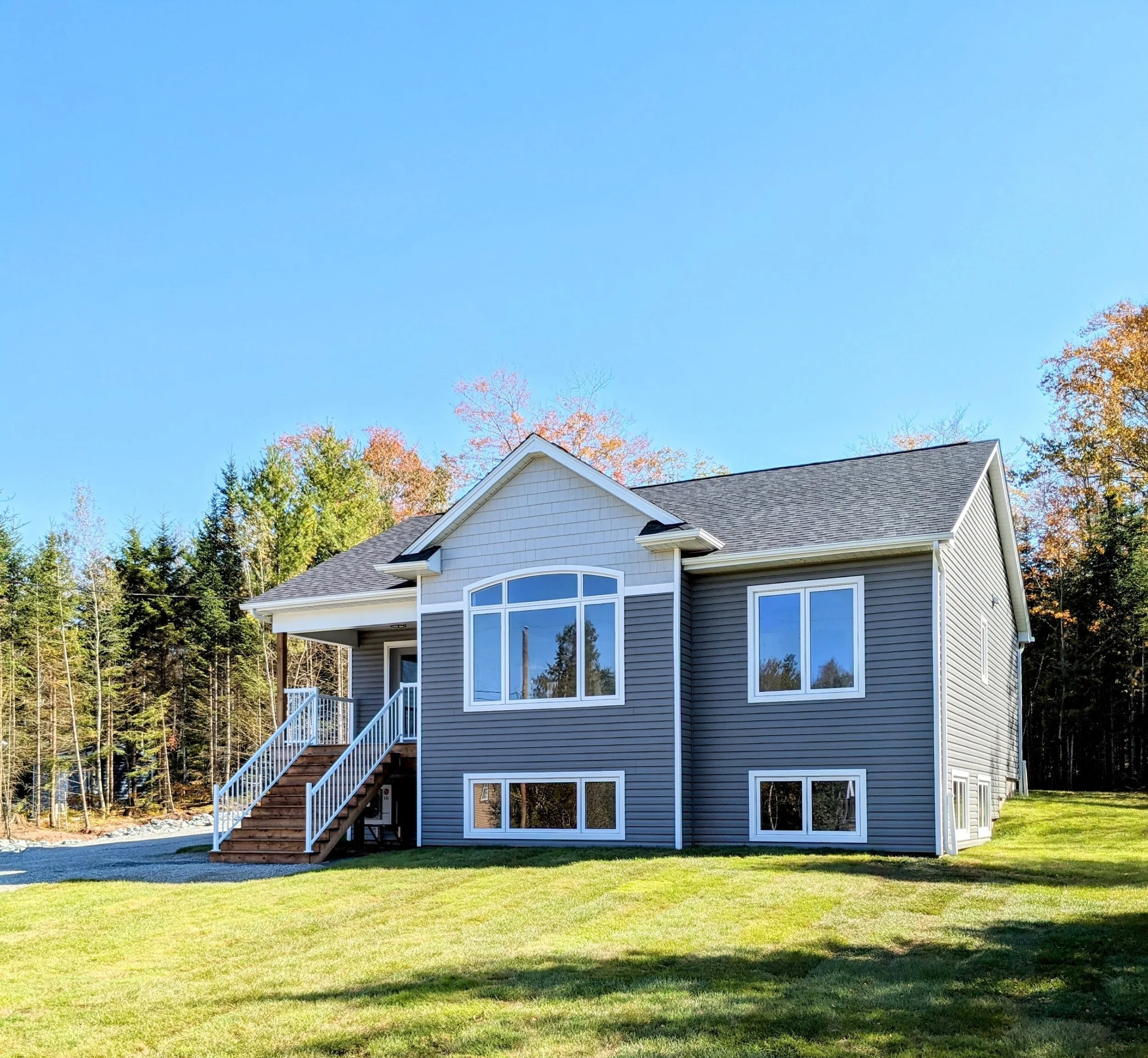 A two-story house with gray siding, a staircase with white railings leading to a front porch, multiple large windows, and a backyard with green grass, surrounded by trees under a clear blue sky.