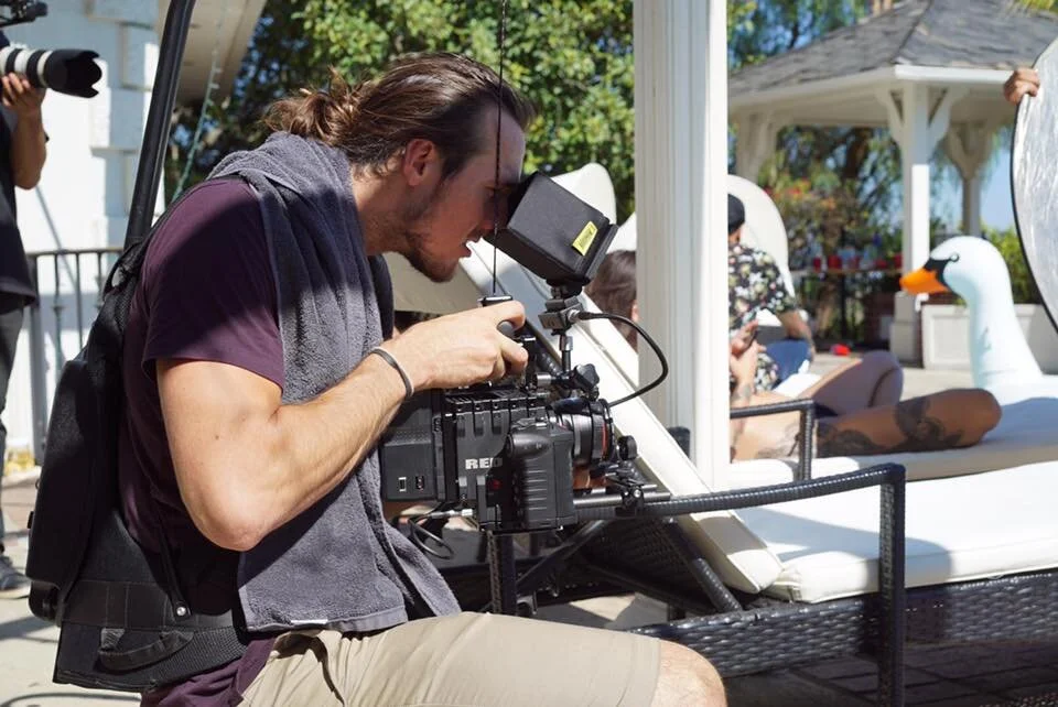 A man filming with a professional camera outdoors near a pool, with a person lounging on a white pool float shaped like a swan in the background.