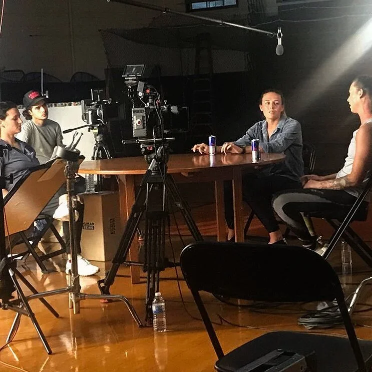 A behind-the-scenes view of a video interview setup with four women at a wooden table, cameras, and lighting equipment in a dimly lit studio.