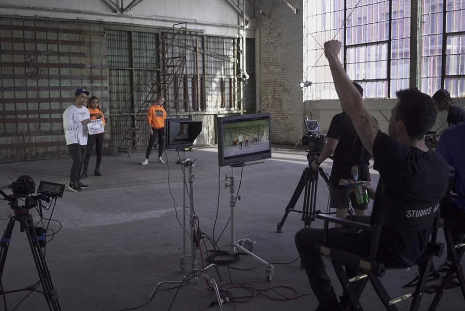 Film crew filming a scene with actors in an industrial warehouse setting, with professional camera equipment and monitors.