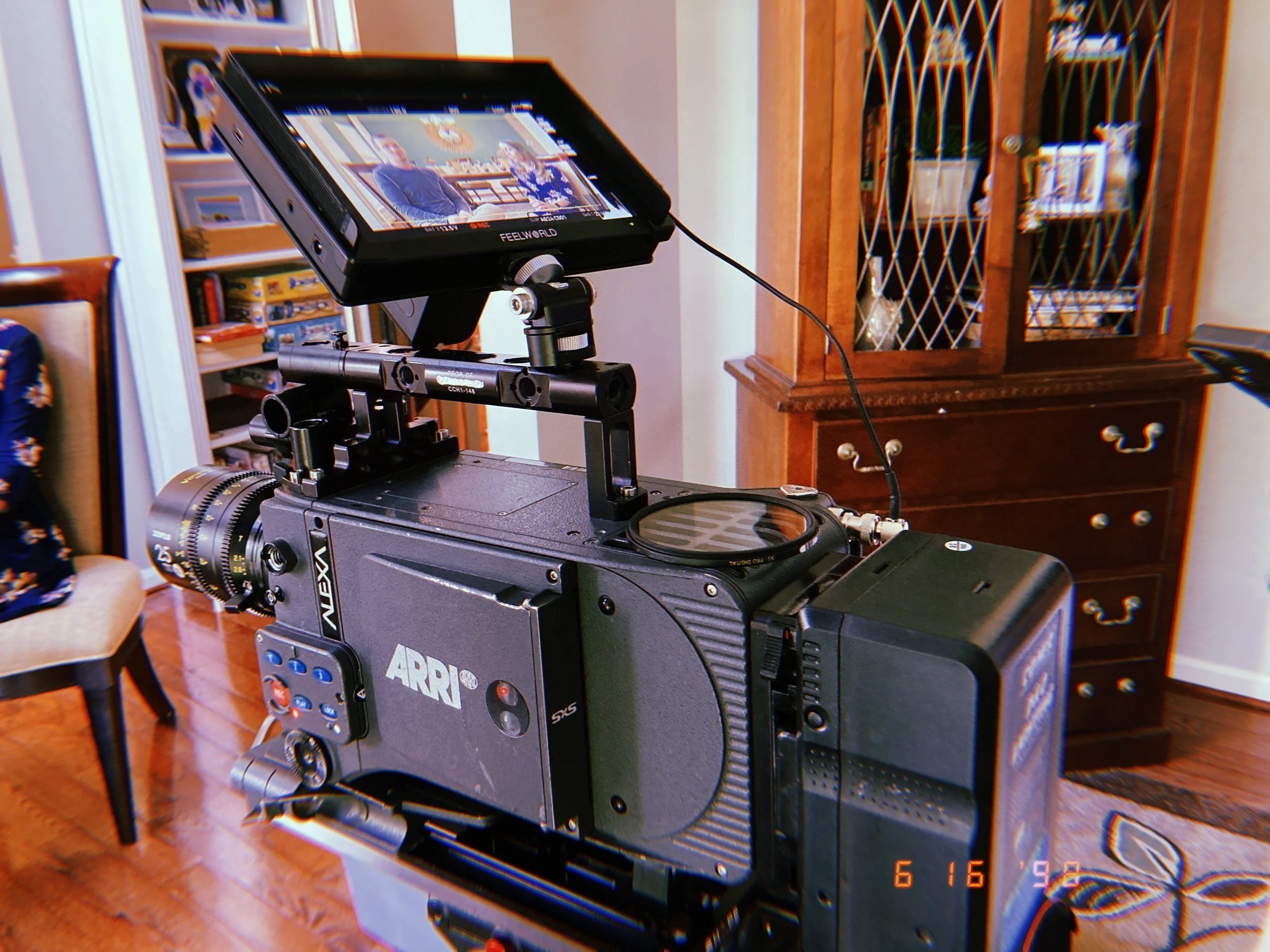 A professional ARRI Alexa camera with a monitor mounted on top is set up indoors, likely for filming or photography, with furniture and bookshelf visible in the background.