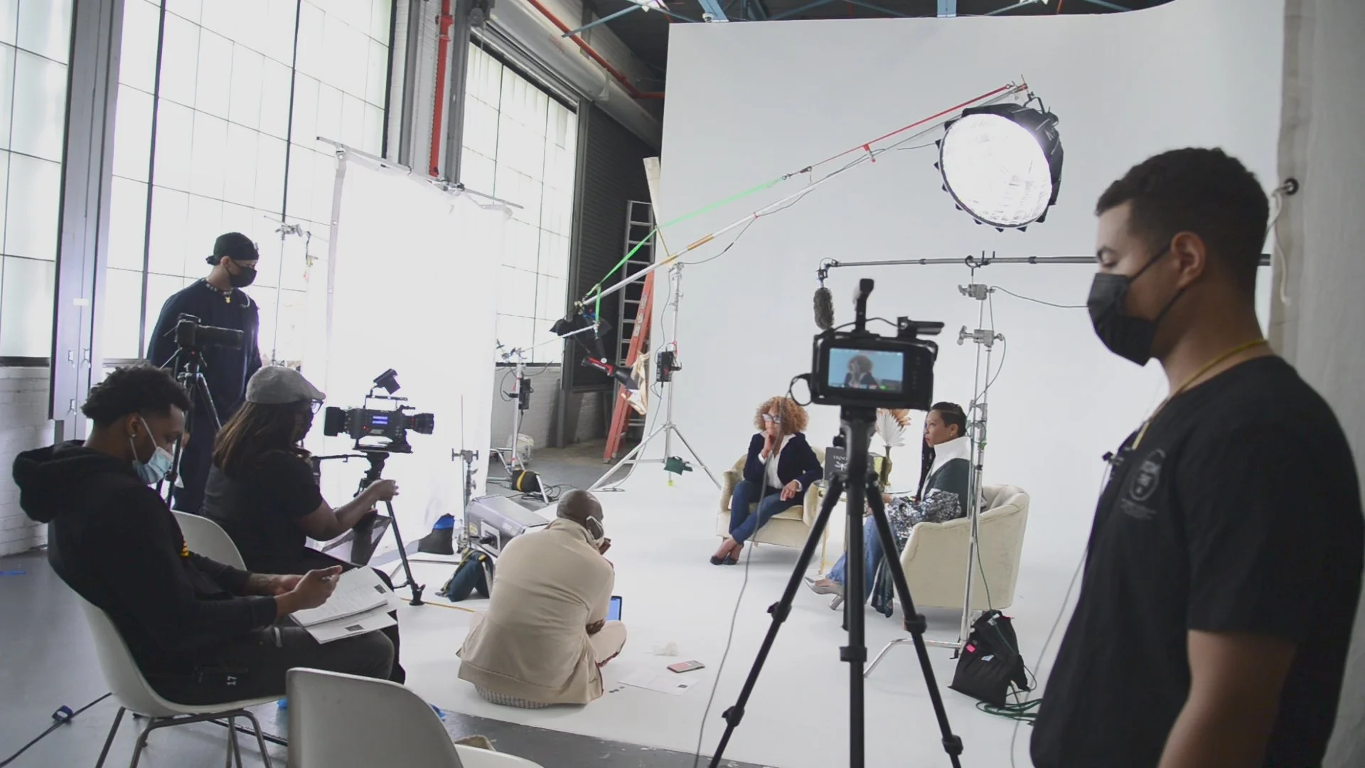 People filming and preparing for a photo shoot or interview in a studio with a white backdrop, professional lighting, and cameras, with some team members reviewing notes and operating equipment.