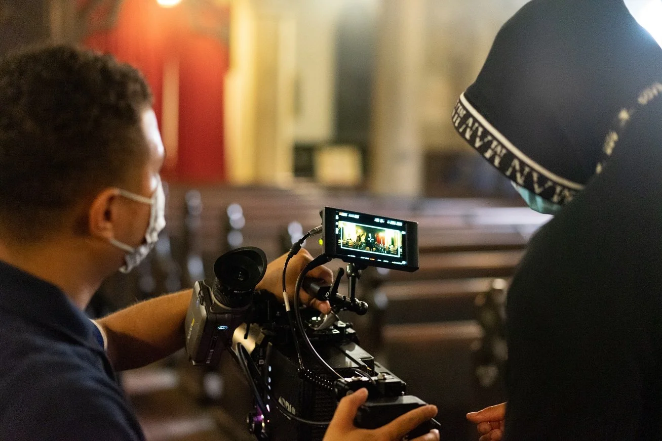 Two men, one operating a professional video camera, the other observing, in a dimly lit indoor space with wooden tables, both wearing face masks.