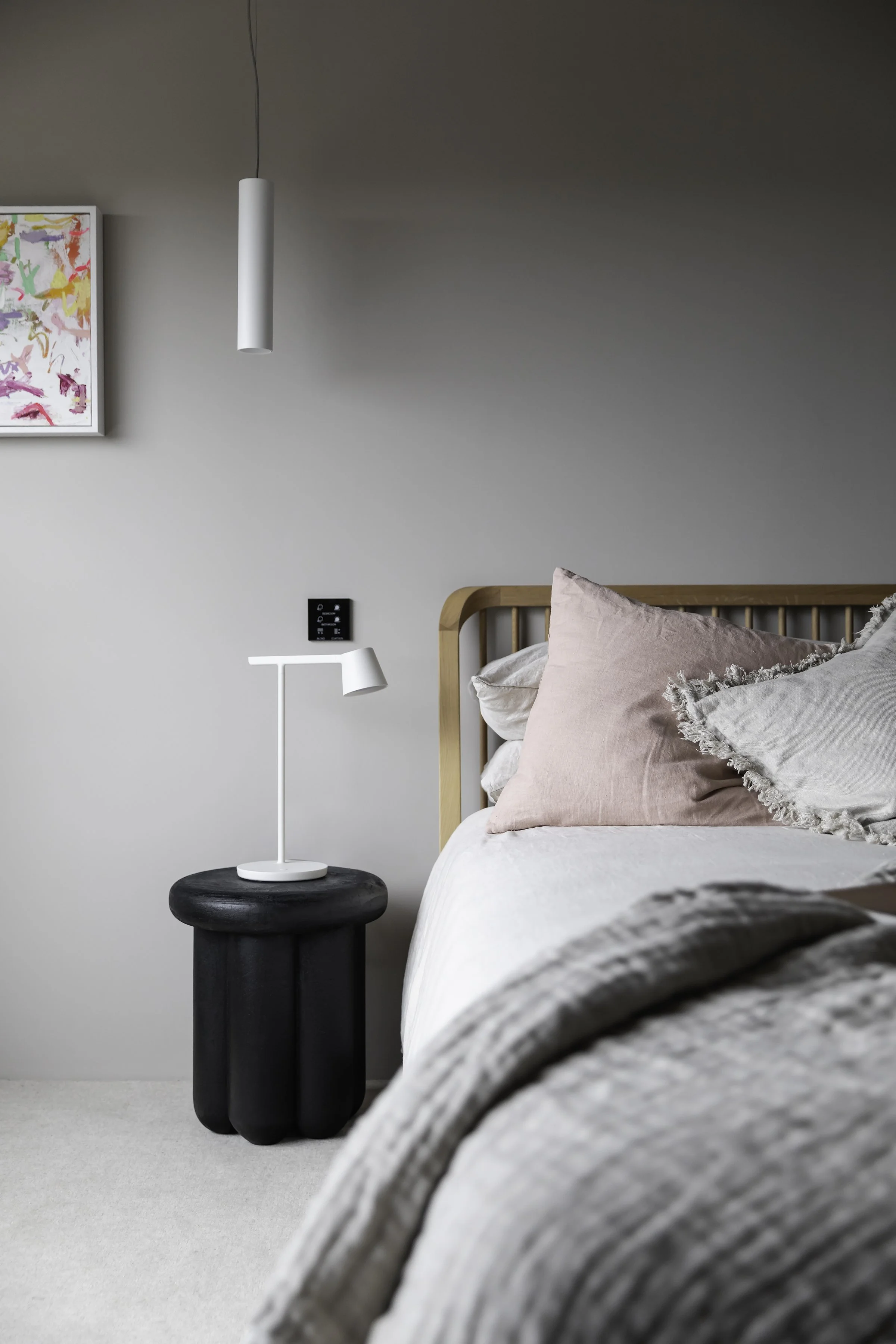 Minimalist bedroom with a bed, light gray bedding, wooden headboard, black side table, white lamp, and wall art.