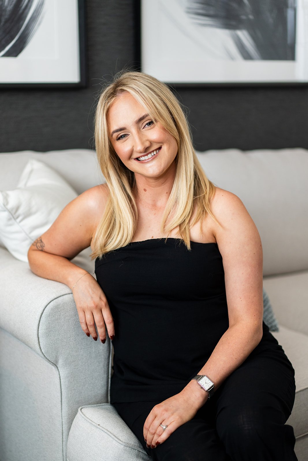 A smiling blonde woman in a black strapless top sitting on a light-colored couch in a modern indoor setting with black and white abstract art on the wall behind her.