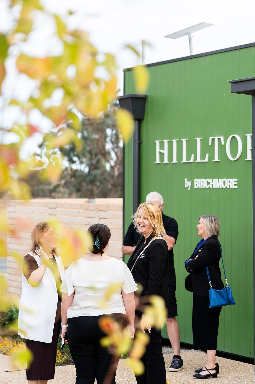 A group of people standing outdoors near a green building with the text 'HILLTOP by BIRCHMORE.' Some people are talking, and the scene includes blurred yellow and green foliage in the foreground.