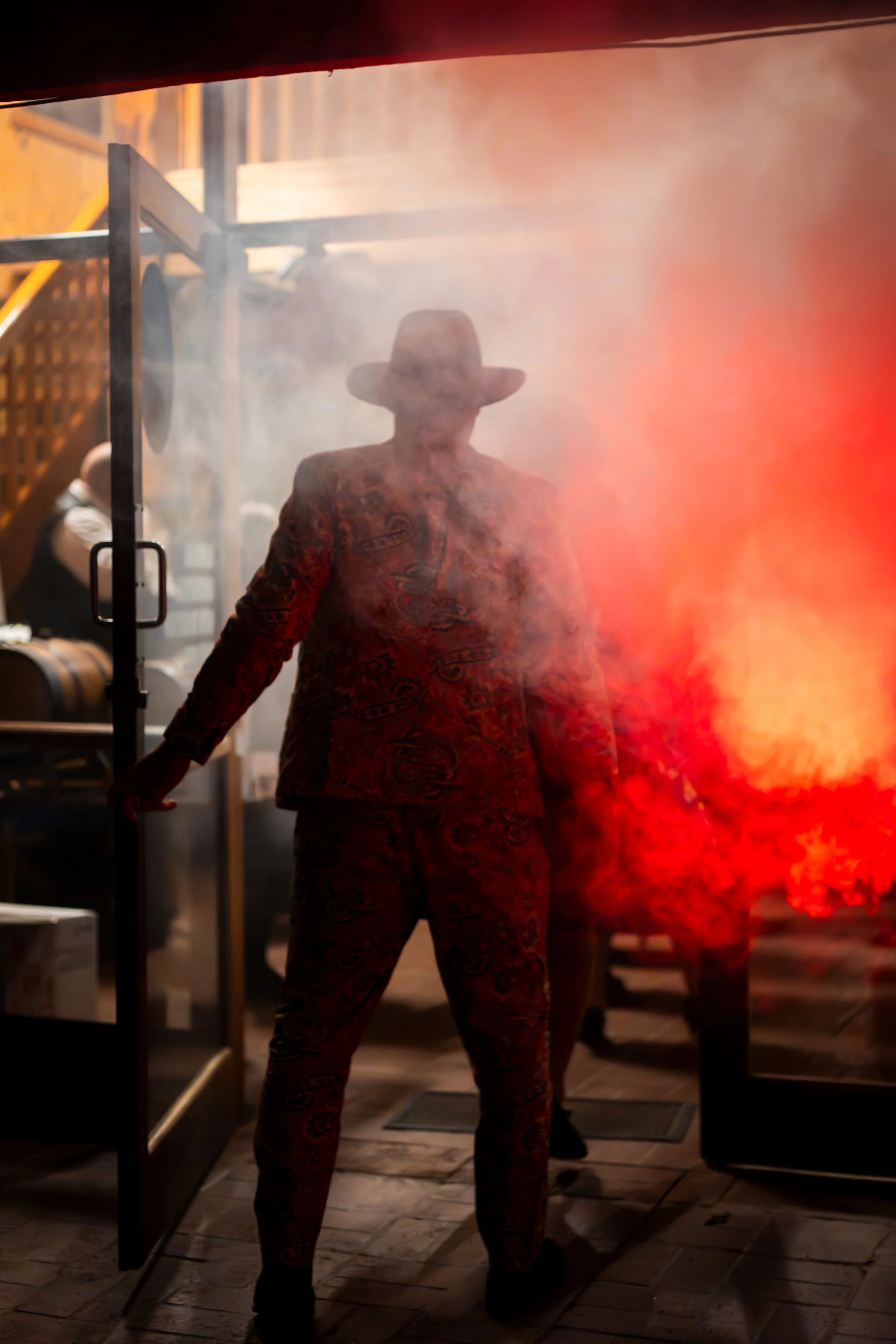 A person wearing a cowboy hat and a patterned outfit stands backlit by red light and smoke, partially obscured, indoors.