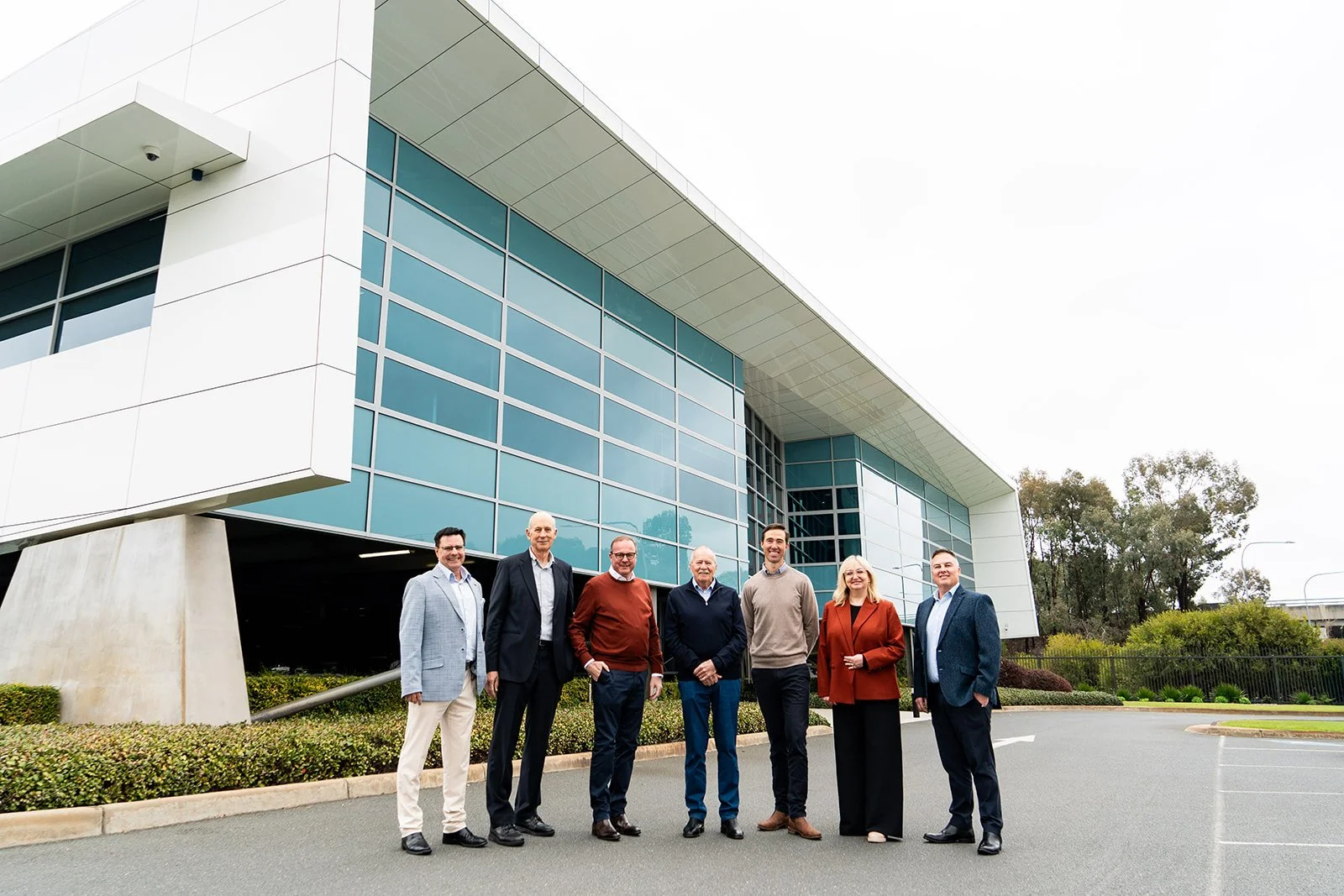 Group of seven people standing outside modern office building with glass windows and curved roof, posing for photo.