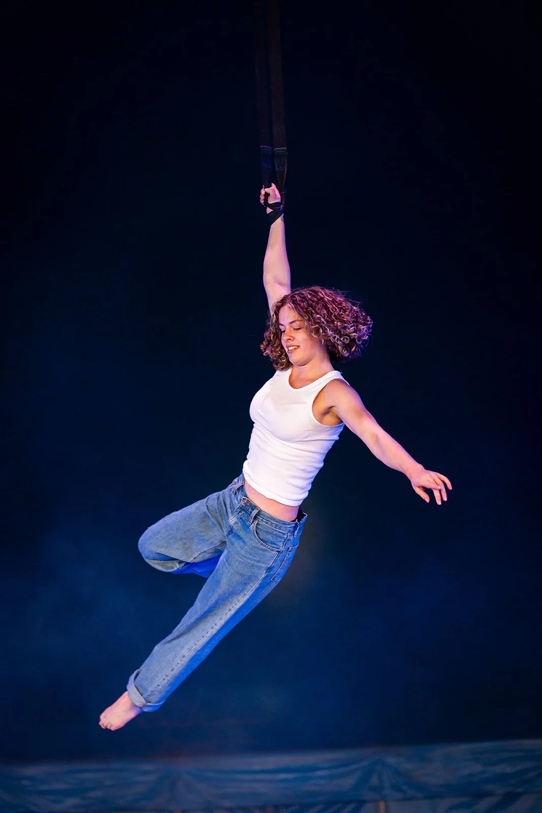 A woman with curly hair in a white sleeveless top and jeans performs aerial acrobatics on a trapeze against a dark background.