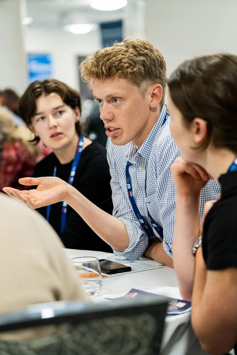 Young man with blond hair in a checkered shirt is speaking to two women at a conference table, with one woman resting her chin on her hand and the other looking on.