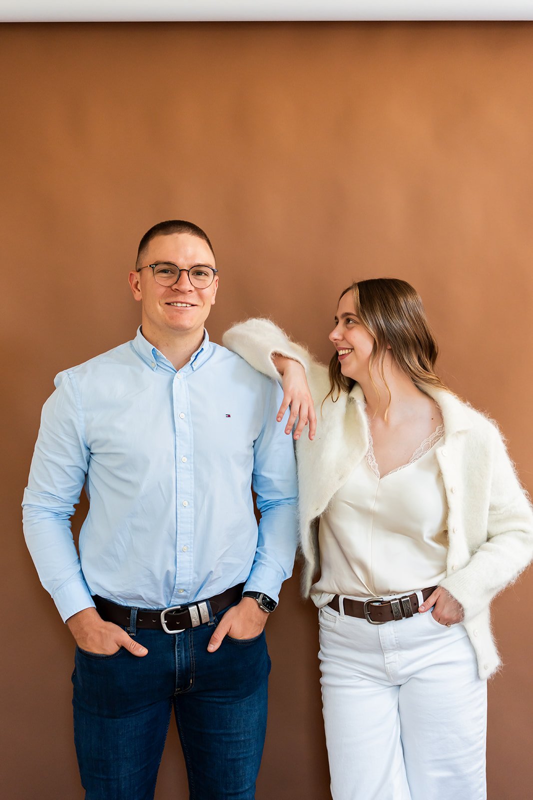 A man and a woman standing together in front of a plain brown background. The man is smiling and wearing glasses, a light blue button-up shirt, dark jeans, and a black belt. The woman is looking at the man, smiling, and wearing a white cardigan over a cream-colored top, white pants, and a brown belt.