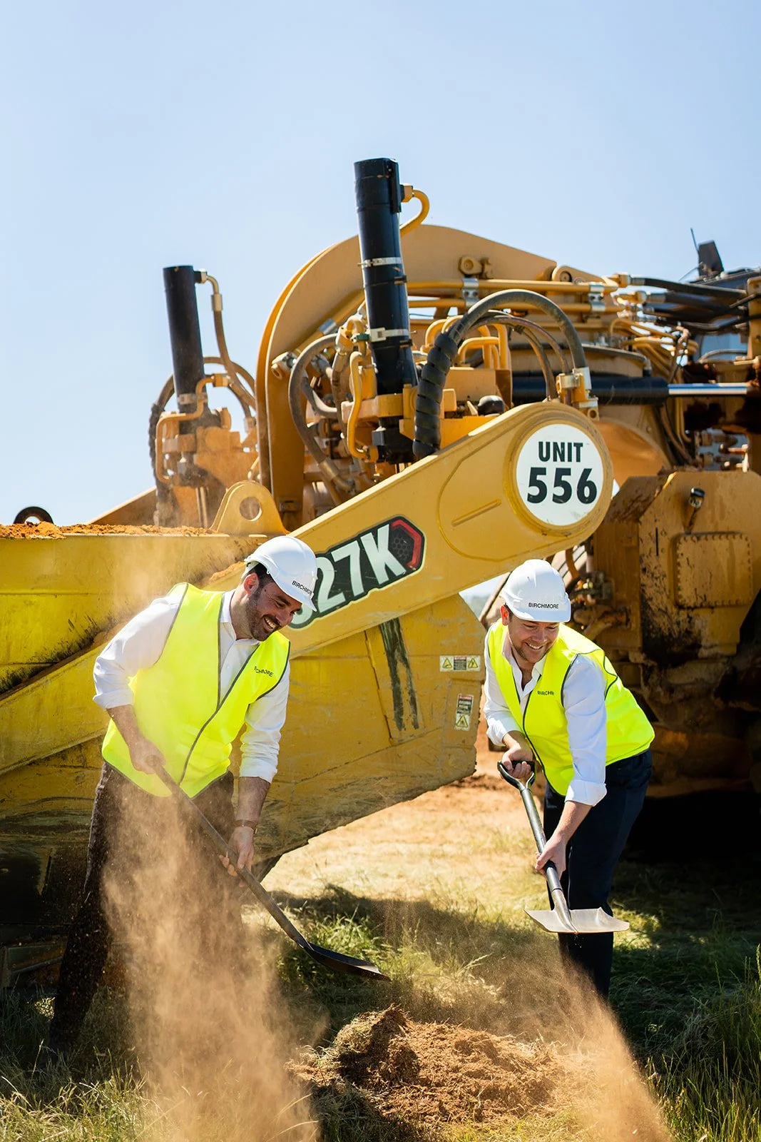 Two construction workers in yellow safety vests and white helmets digging in the ground with shovels in front of a large yellow Caterpillar bulldozer.