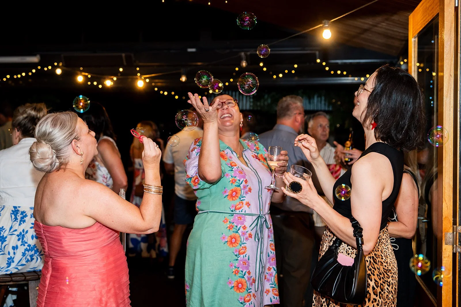 Women celebrating with bubbles at a festive gathering, some holding glasses of wine, with string lights overhead and other guests in the background.