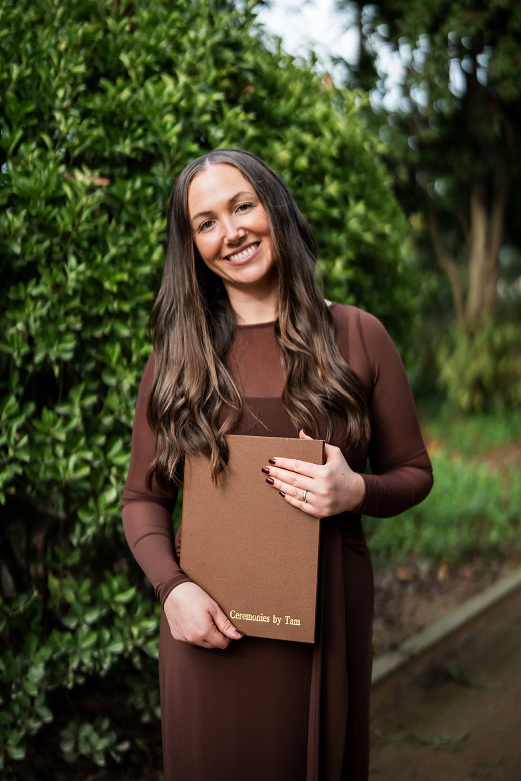 A smiling woman with long brown hair in a brown dress holding a brown folder labeled 'Ceremonies by Tam' outdoors with green bushes and trees in the background.