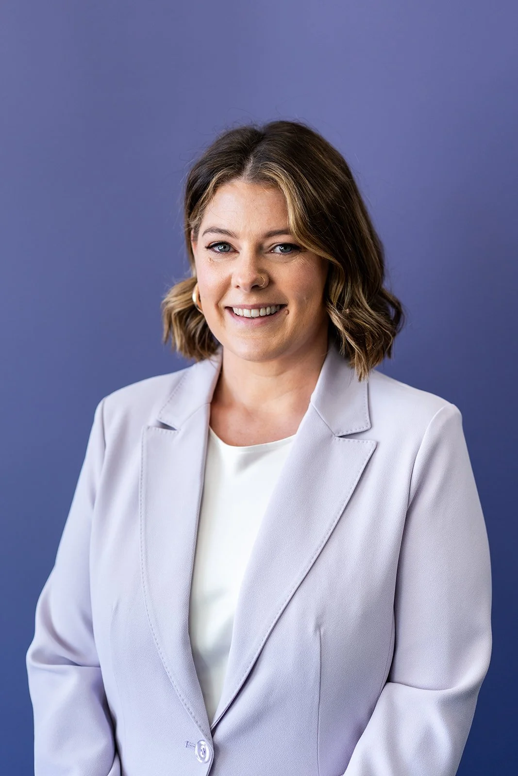 A professional woman with shoulder-length brown hair, wearing a light gray blazer and white top, smiling against a blue background.
