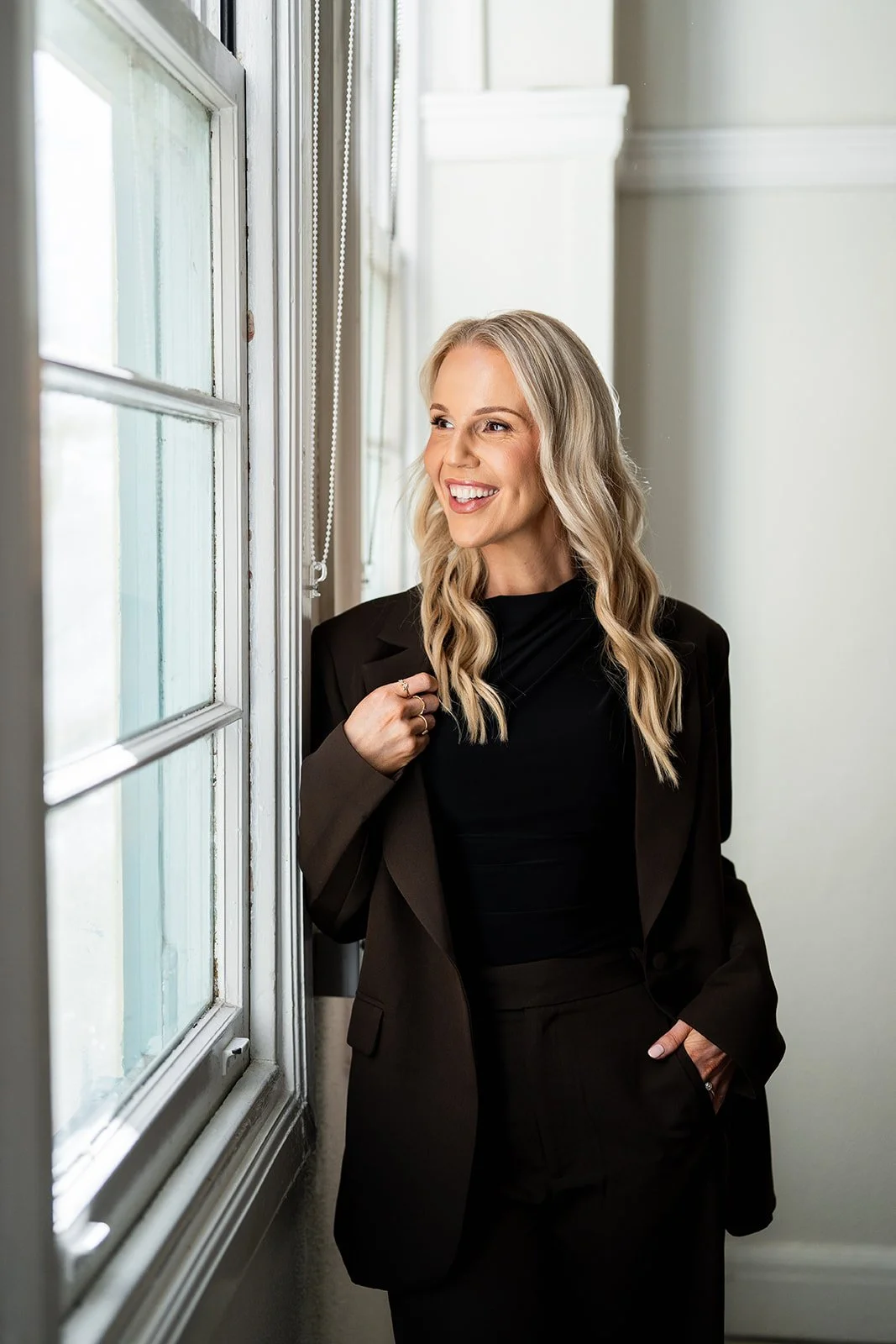 A woman with long blond wavy hair and fair skin, dressed in a black blazer and black top, smiling and looking out a window.