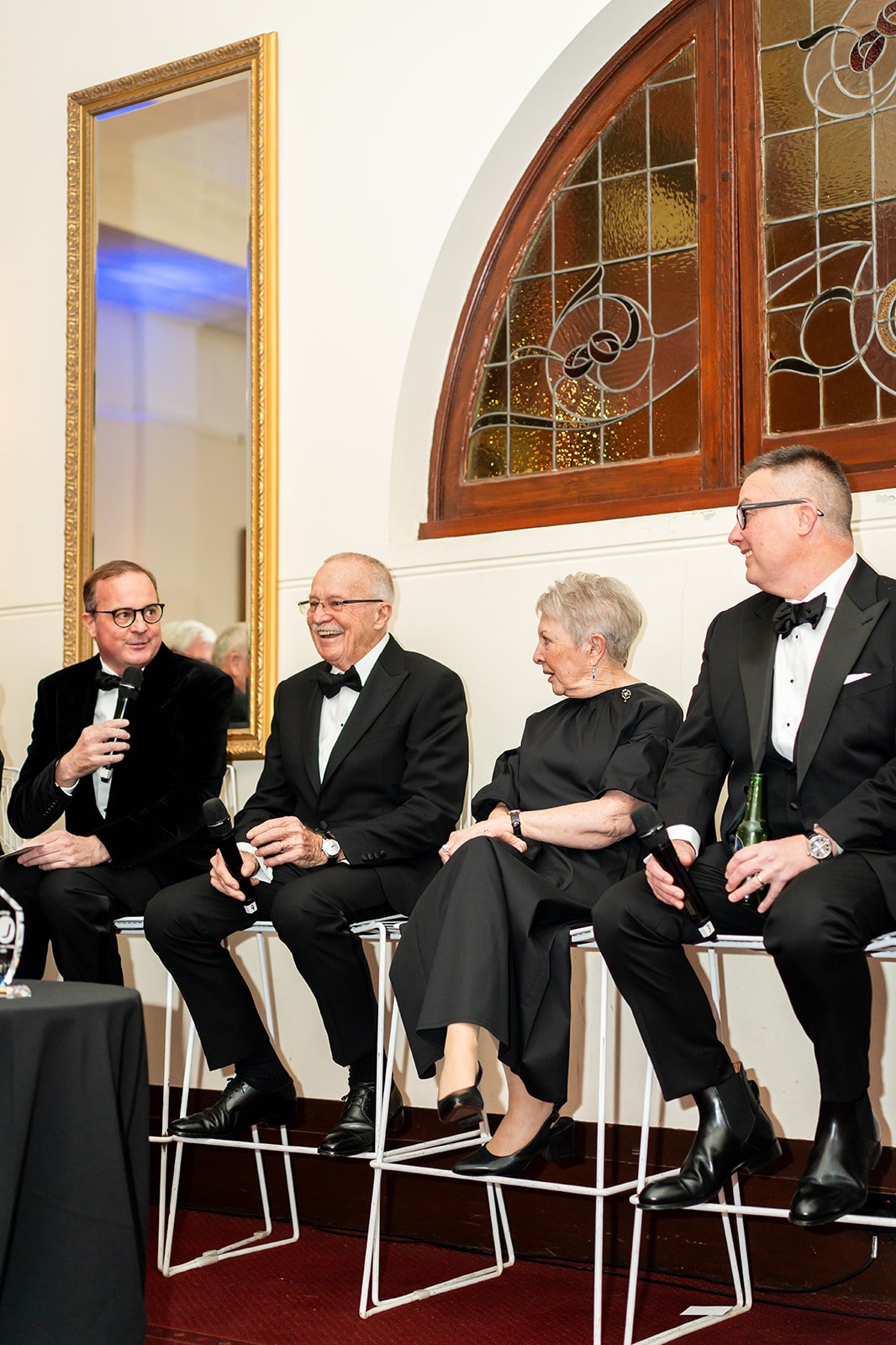 Group of five people in formal attire sitting on a high bench at a formal event. They are smiling and holding microphones. A large mirror and stained glass window are in the background.