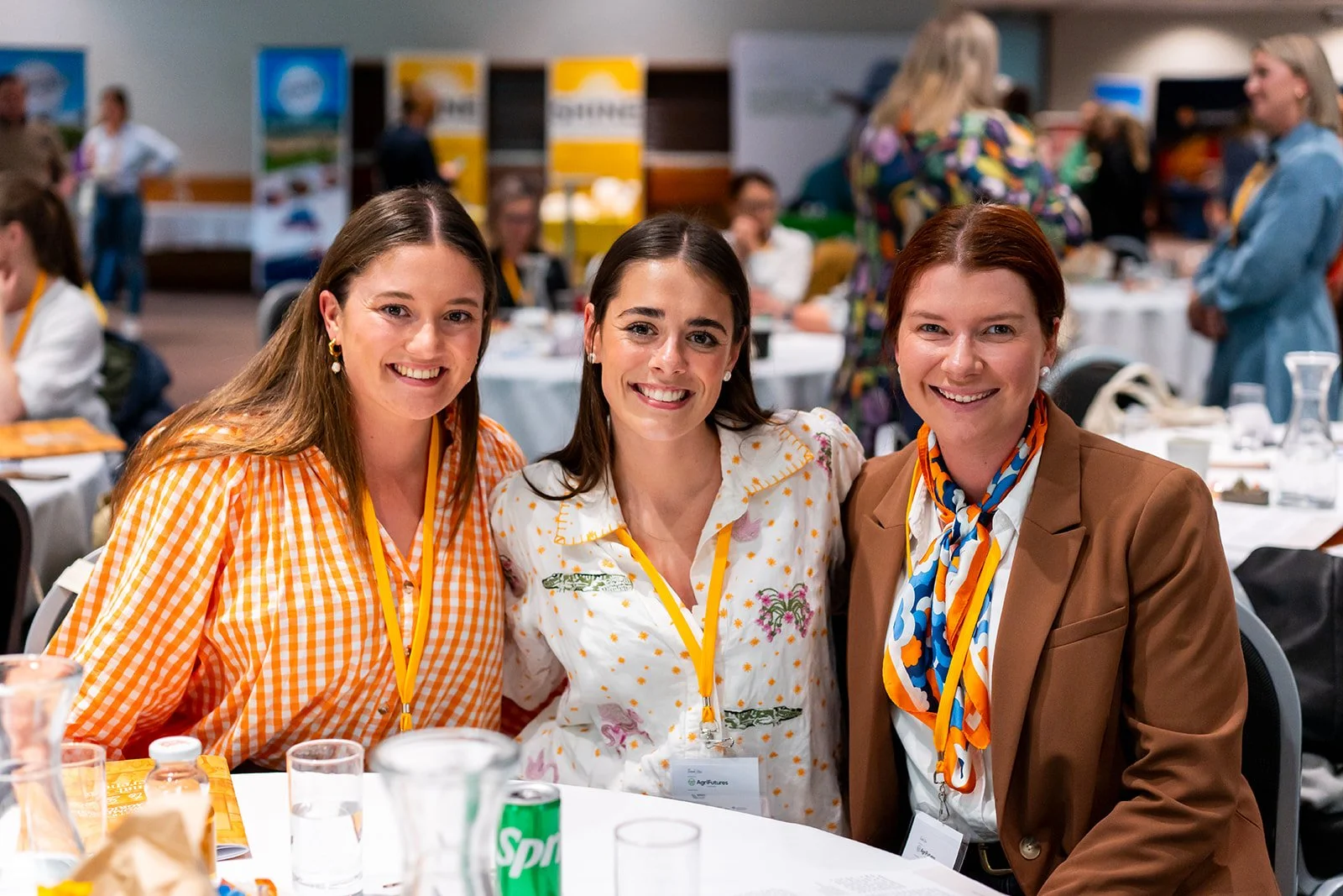 Three women smiling and sitting together at a conference or event, with a background of other attendees and tables.
