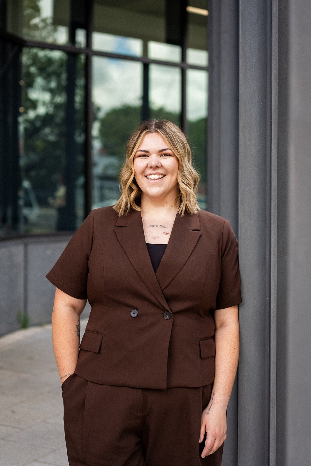 A woman with shoulder-length blonde hair, smiling, wearing a brown suit with short sleeves, standing outdoors near a modern building with large windows.