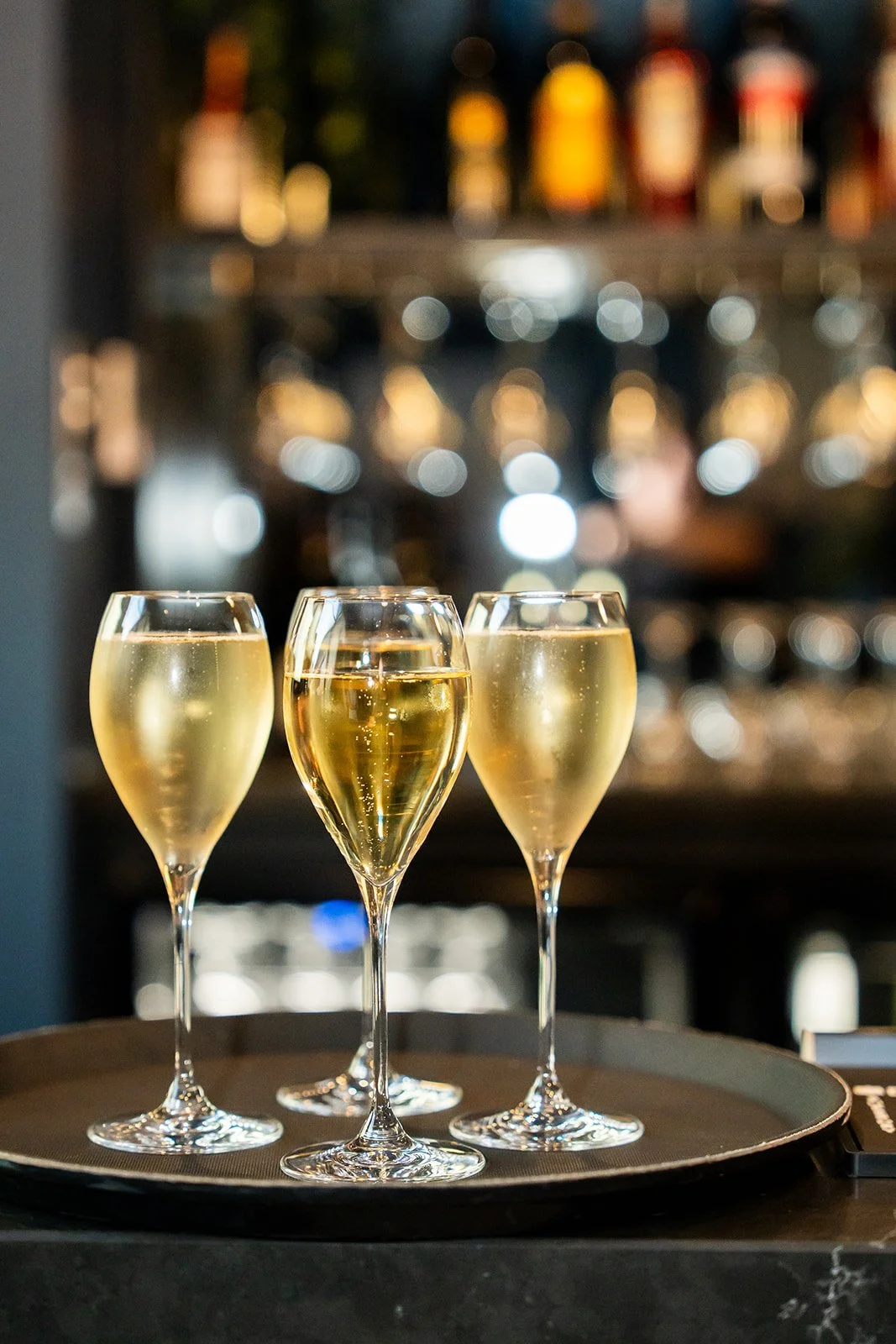 Three glasses of champagne on a tray, with a blurred bar background.