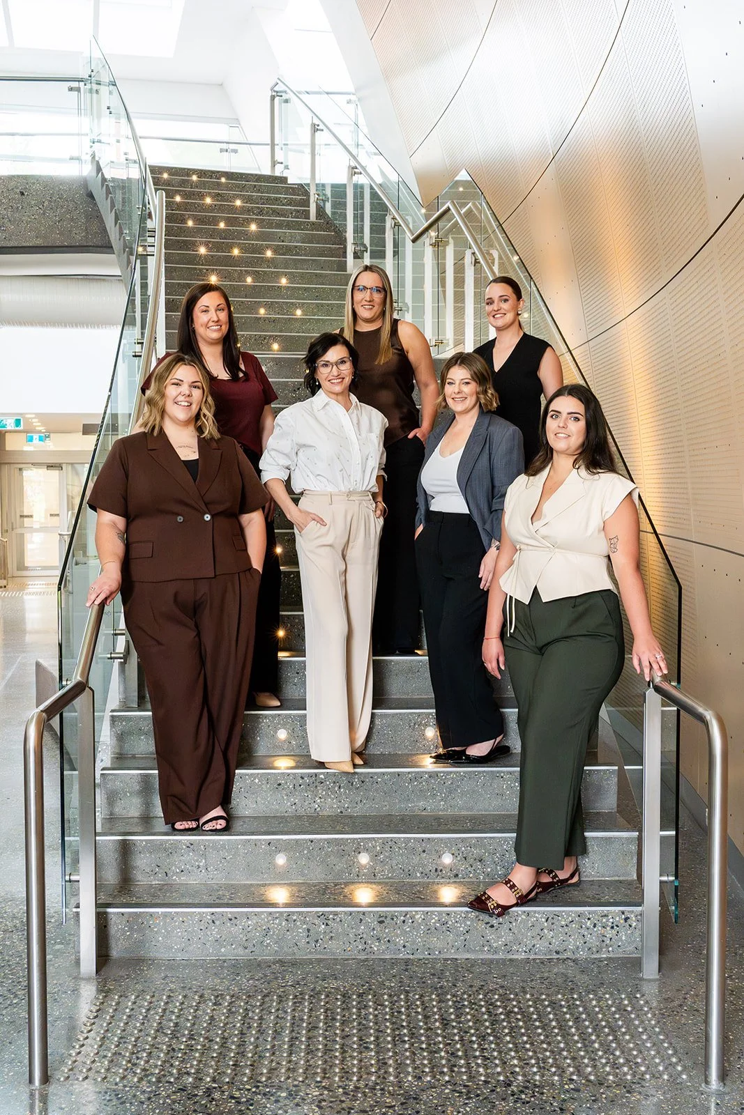 Group of eight women standing on a staircase inside a modern building, smiling and dressed in business casual attire.